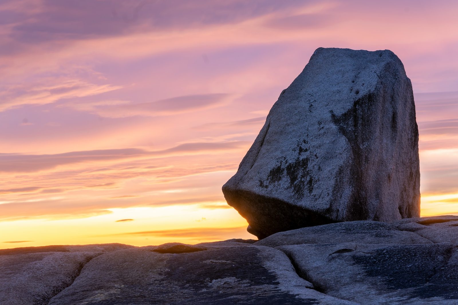 A large granite boulder balances on a rock platform beneath a pink and orange dusk sky.