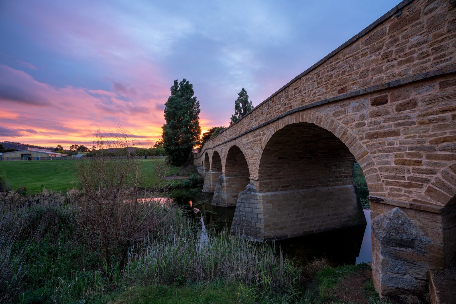 A sandstone arch bridge spans a still river at sunset, its multiple arches reflected in the water below.