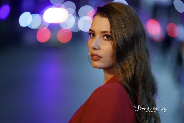 A woman in red looks back over her shoulder on a night street, city lights thrown out of focus behind her