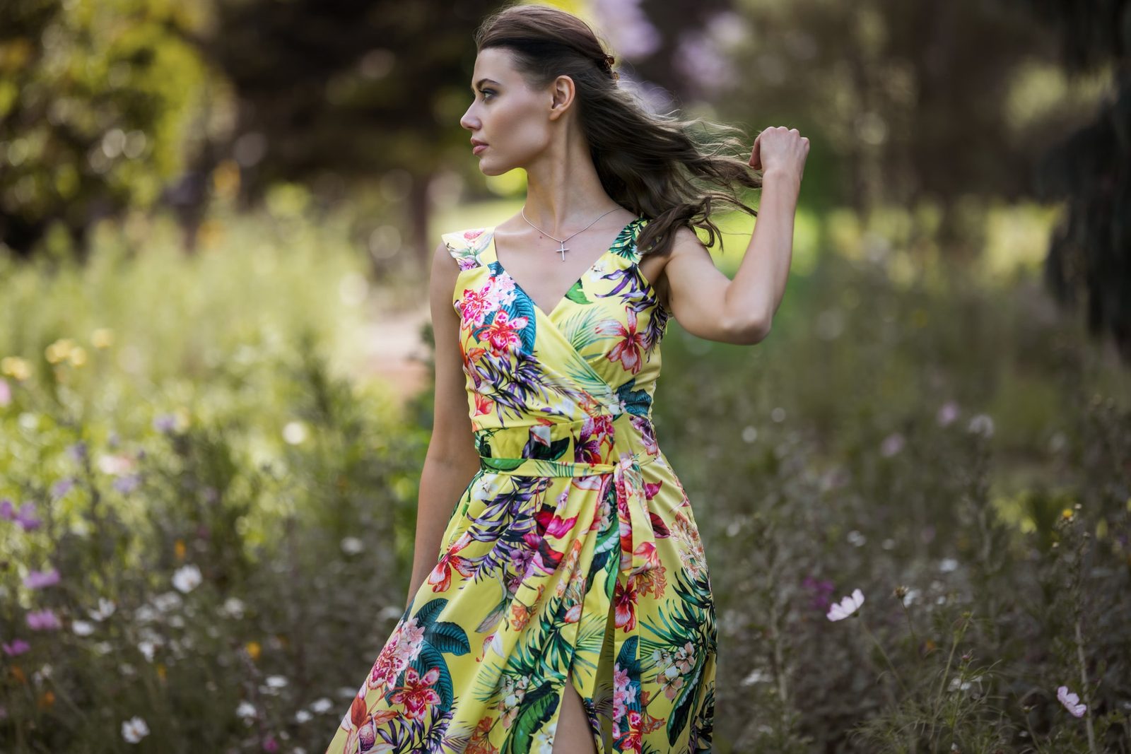 A woman in a yellow tropical-print wrap dress walks through a wildflower meadow, hair lifting in the breeze as she looks aside
