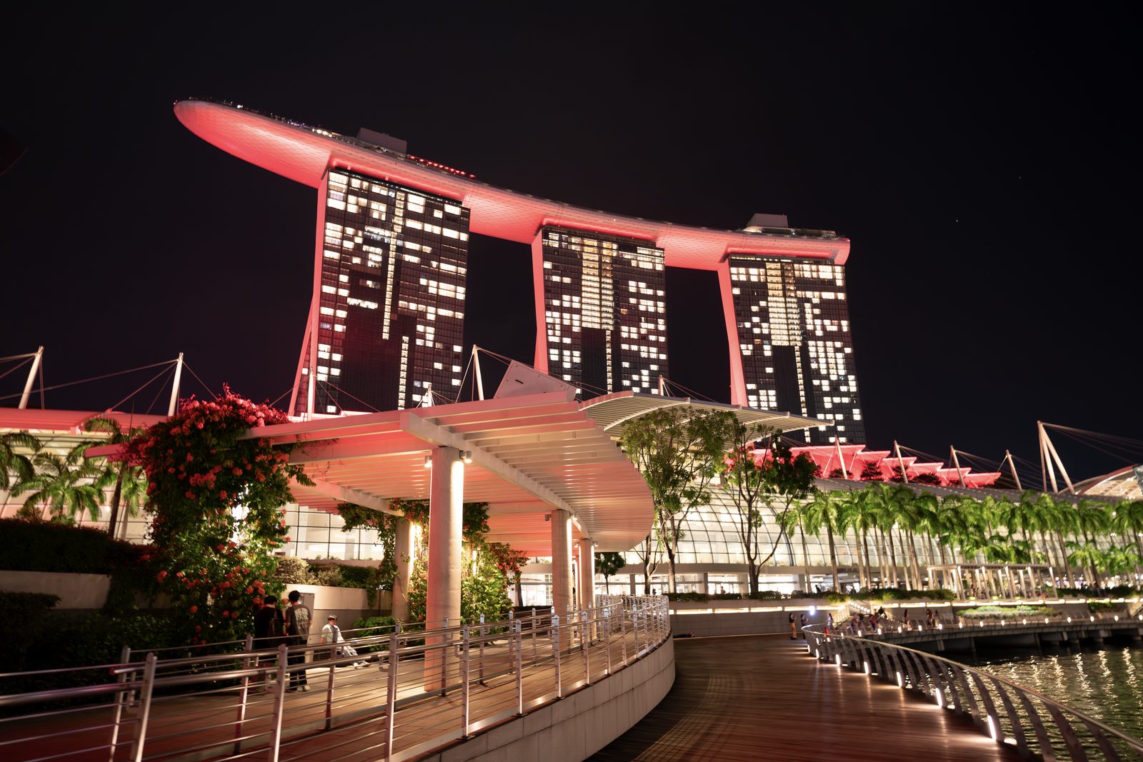 The three towers of a hotel are lit red above a waterfront boardwalk, bougainvillea and palms along the rail.