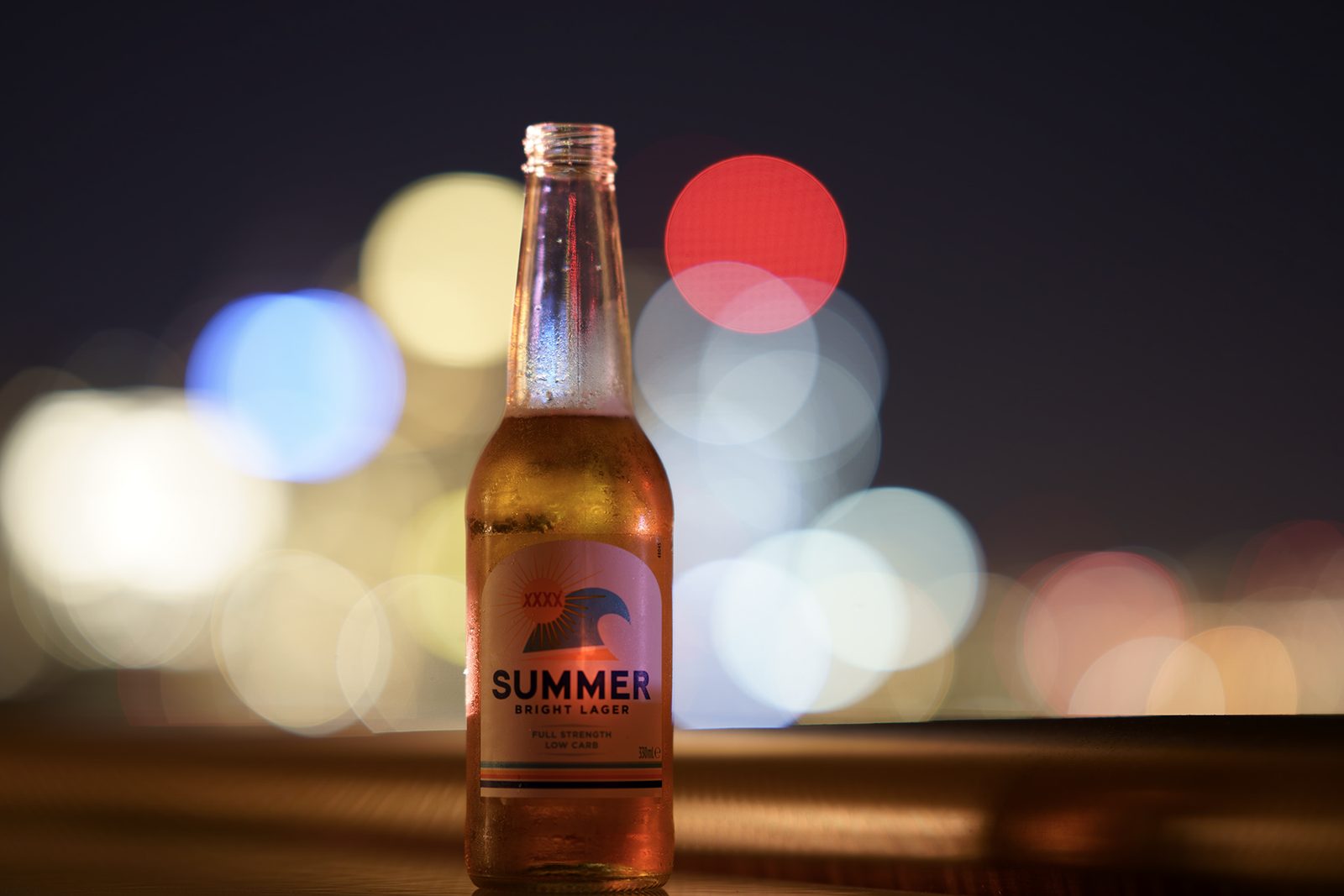 A cold beer bottle stands on a timber rail against a wall of out-of-focus city lights at night.