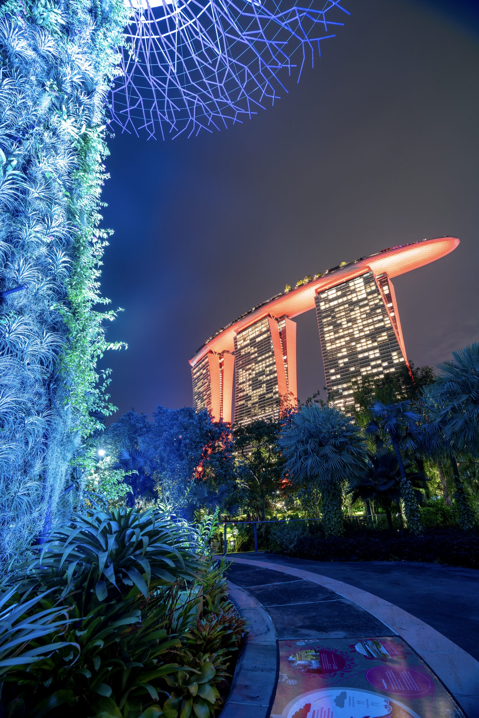 A supertree trunk glows blue in the foreground while a three-towered hotel stands lit orange beyond the palms.