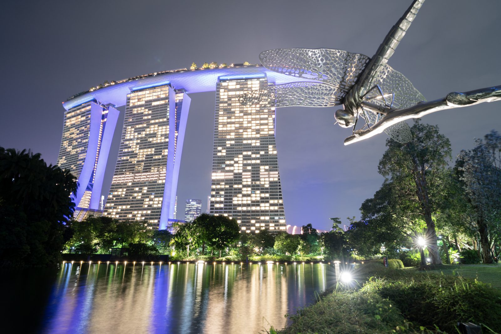 A large metal dragonfly sculpture perches in the foreground while three lit hotel towers stand reflected in still water.