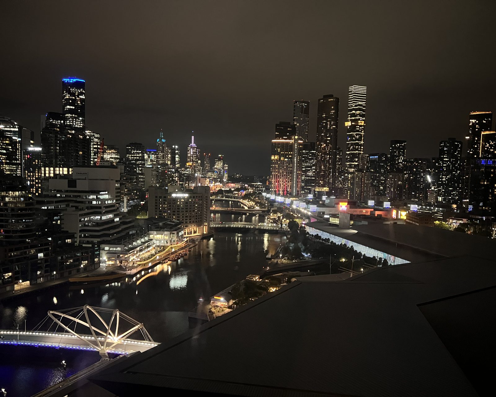 Melbourne's towers and the Yarra seen from above at night, a lit pedestrian bridge crossing the dark water below.