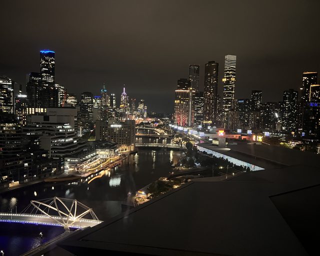Melbourne's towers and the Yarra seen from above at night, a lit pedestrian bridge crossing the dark water below.