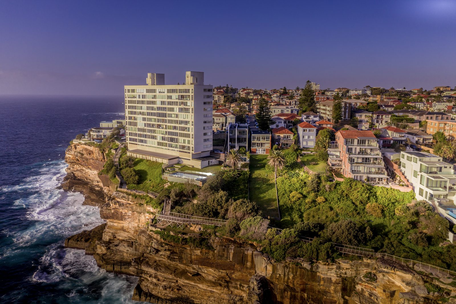 An apartment block and houses crowd a sandstone clifftop above breaking surf, seen from the air in low sun.