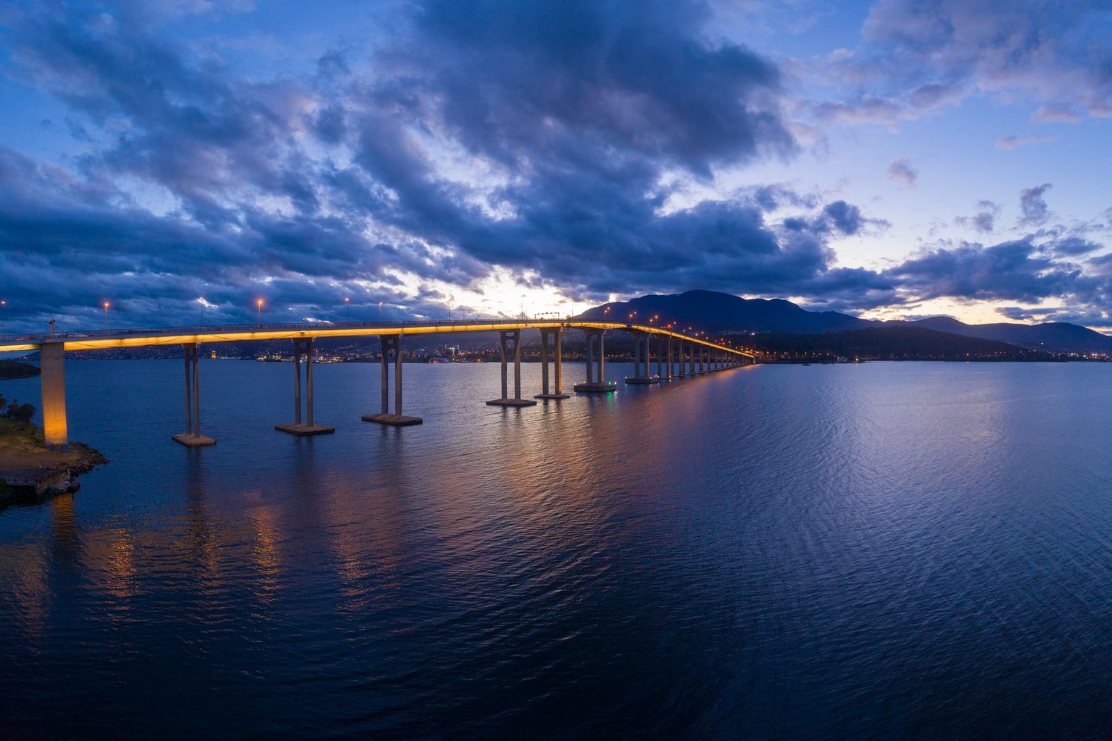 A long causeway bridge lit gold at dusk stretches across still water toward a mountain silhouette under heavy cloud.