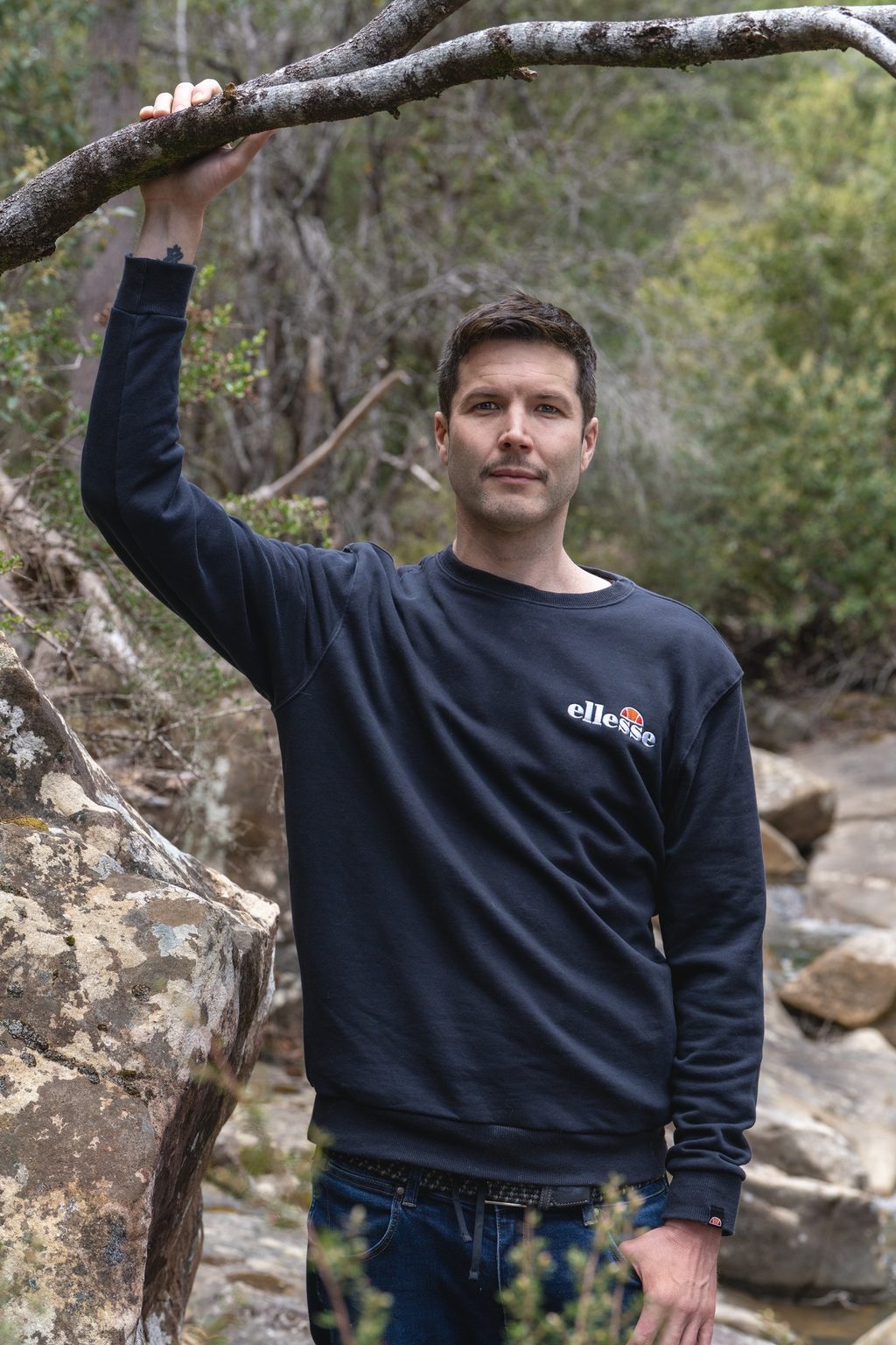 Tim Downey standing in bushland beside a rocky creek, one hand resting on a low bare branch overhead, wearing a navy sweatshirt